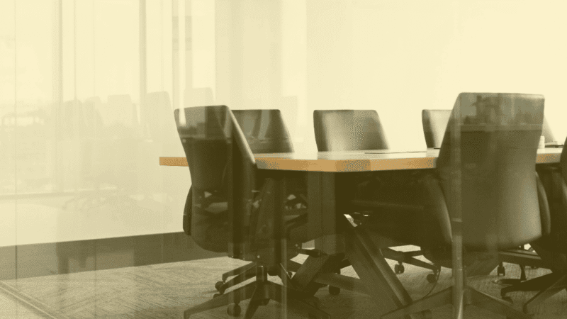 Empty conference room with black office chairs around a wooden meeting table seen through a glass wall.